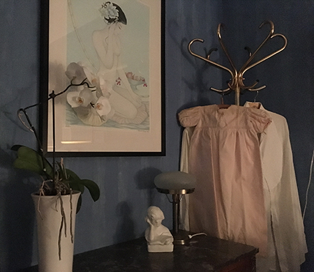 Detaljer från sovrummet: en orkidé, en tavla, ett vitt bokstöd, en lampa, en rosa dopklänning och två frackskjortor.