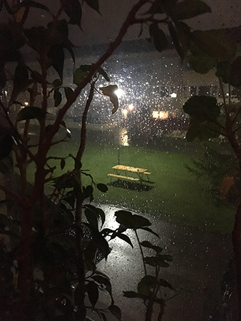 Regnig morgon genom köksfönstret. En träbänk med bord är upplyst av en lampa.