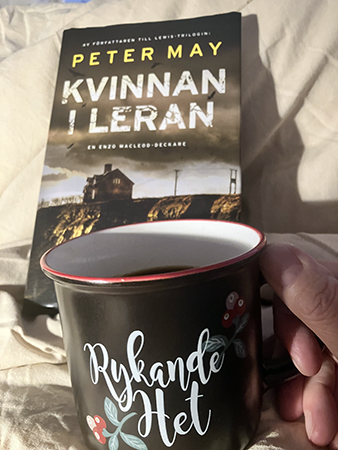Rykande het mugg kaffe på sängen o boken Kvinnan i leran