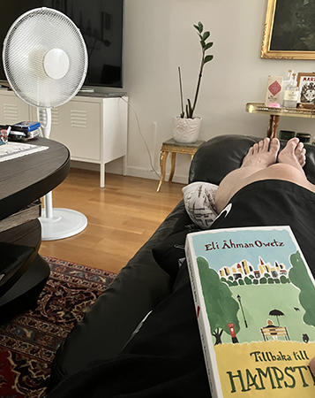 Läser Tillbaka till Hampstead iförd shorts i soffan med golvfläkten på